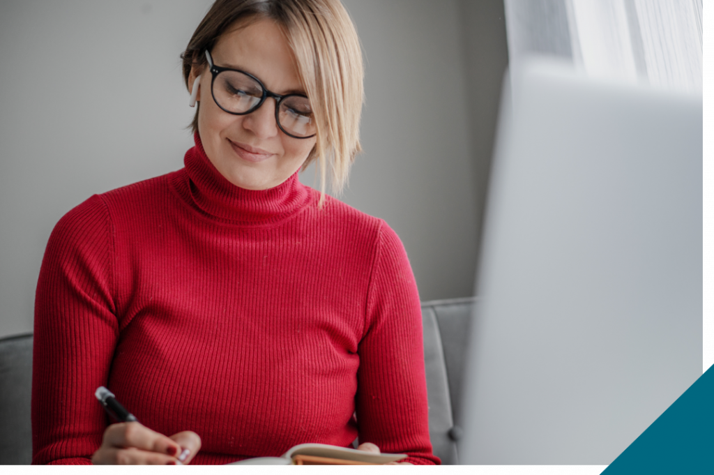 Woman writing in notebook with laptop open.